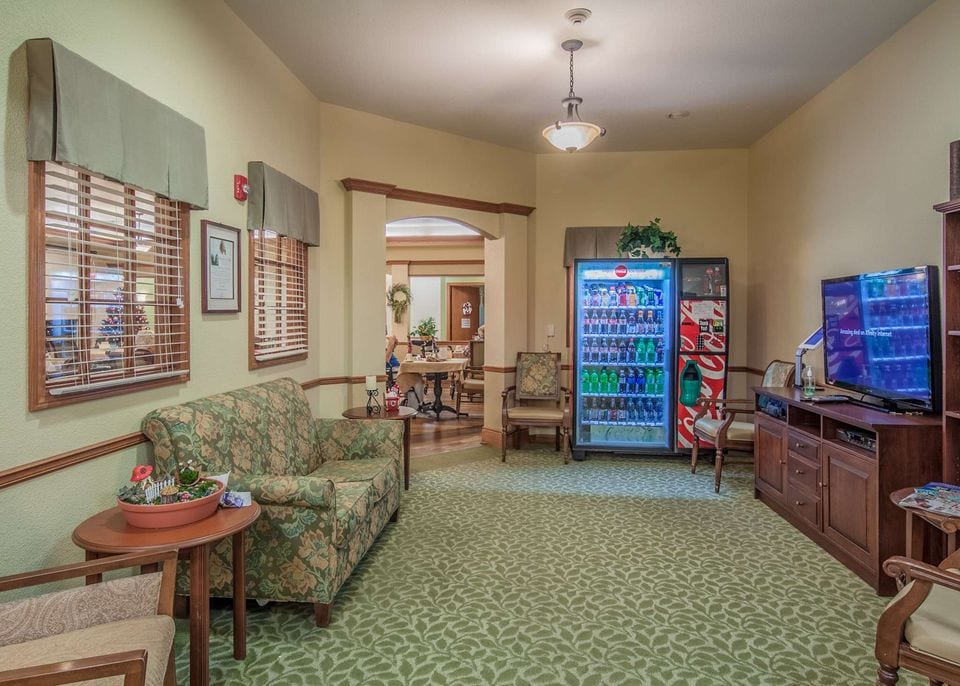 A lounge with a floral sofa, wooden-armed chairs, a TV cabinet and a drinks vending machine